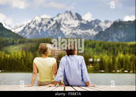 Senior paar sitzt am Pier über dem Bergsee mit den Bergen im Hintergrund Stockfoto