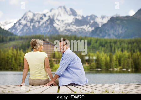 Senior paar sitzt am Pier über dem Bergsee mit den Bergen im Hintergrund Stockfoto