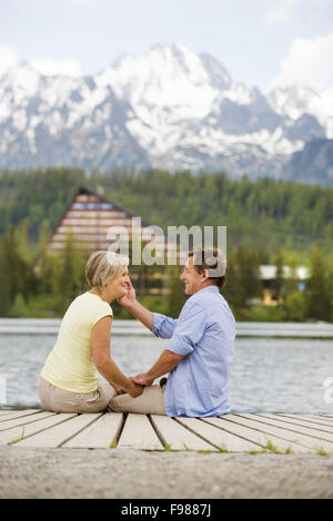 Senior paar sitzt am Pier über dem Bergsee mit den Bergen im Hintergrund Stockfoto