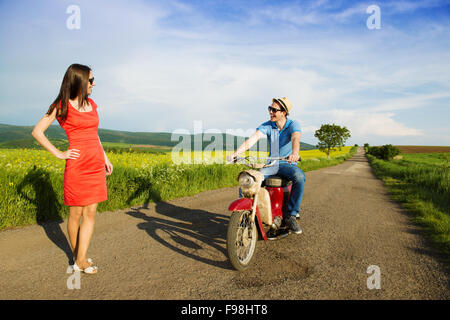 Schöne Mädchen in rotem Kleid ist glücklicher junger Mann auf Retro-Motorrad Abholung. Stockfoto