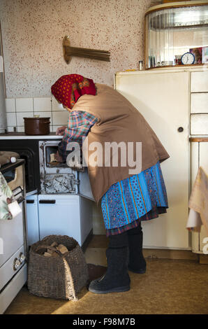 Sehr alte Frau setzt Holz in ihrem alten Holzofen in ihrer Landhausküche Stockfoto