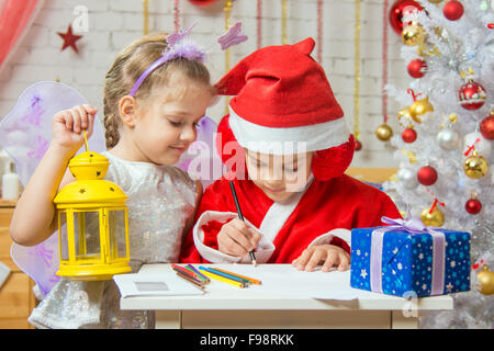 Eine Mädchen, gekleidet in einen roten Anzug Weihnachtsmann zieht Bleistifte sitzen am Tisch, ist als nächstes ein Mädchen gekleidet wie eine Fee mit der candlest Stockfoto