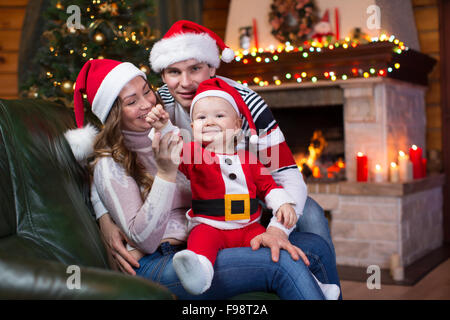 Glückliche Familie mit Kind weared Santa sitzt auf dem Sofa vor dem Weihnachtsbaum und Kamin im Wohnzimmer Stockfoto