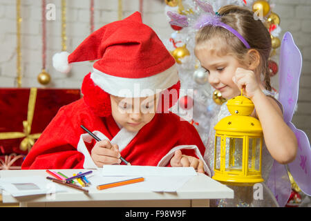 Eine Mädchen, gekleidet in einen roten Anzug Weihnachtsmann zieht Bleistifte sitzen am Tisch, ist als nächstes ein Mädchen gekleidet wie eine Fee mit der candlest Stockfoto