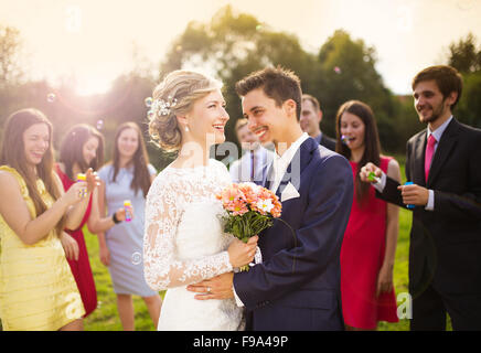 Junge Brautpaar genießen romantische Momente zusammen auf Hochzeit außerhalb, Gäste Hochzeit im Hintergrund Seifenblasen Stockfoto