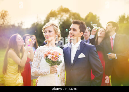 Junge Brautpaar genießen romantische Momente zusammen auf Hochzeit außerhalb, Gäste Hochzeit im Hintergrund Seifenblasen Stockfoto