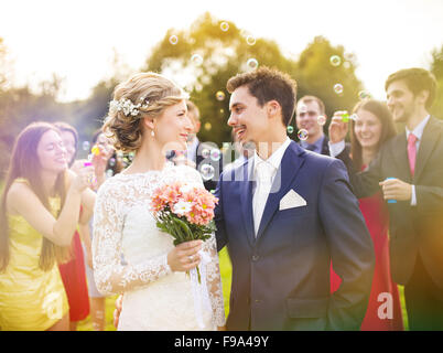 Junge Brautpaar genießen romantische Momente zusammen auf Hochzeit außerhalb, Gäste Hochzeit im Hintergrund Seifenblasen Stockfoto