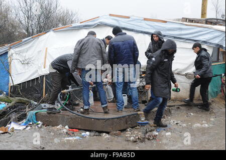Blick auf die Hälfte gebaut aus Holz Unterschlupf in den überfüllten, schlammigen Bedingungen im Dschungel Flüchtlingslager, Calais. Das Camp ist Heimat von rund 6.000 Menschen und Bedingungen verschlechtern mit dem nassen Wetter. Ansicht von Flüchtlingen sammeln von Wasser aus einem Hahn Stand Rohr. Stockfoto