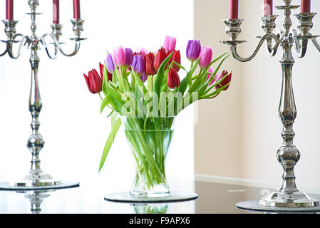 Ein klares Glasvase mit Umzug, rot und rosa Tulpen auf einem Glastisch mit zwei Silberleuchter Stockfoto