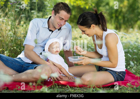 Glückliche junge Eltern füttern ihre Tochter beim Picknick. Stockfoto