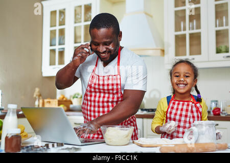 Afrikanisch-amerikanischer Mann Vernetzung und sprechen auf dem Handy während seiner Tochter machen Gebäck aus Teig in der Nähe von Stockfoto