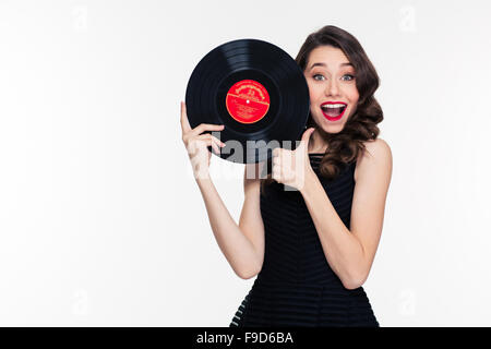 Schöne positive lockiges junge Frau mit Retro-Frisur hält Vinyl-Schallplatte und Daumen über weißem Hintergrund auftauchen Stockfoto