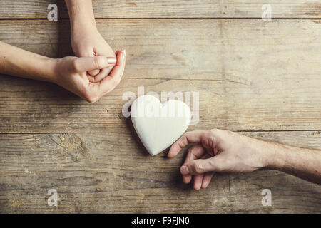 Man bietet ein weißes Herz zärtlich zu einer Frau. Studio auf einem hölzernen Hintergrund erschossen, Ansicht von oben. Stockfoto