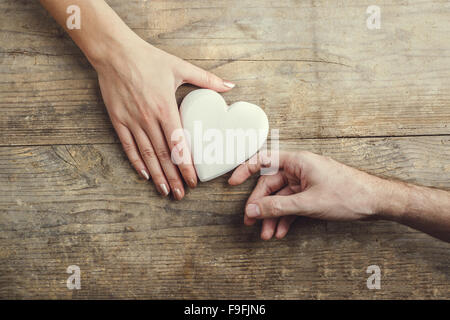 Man bietet ein weißes Herz zärtlich zu einer Frau. Studio auf einem hölzernen Hintergrund erschossen, Ansicht von oben. Stockfoto