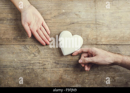 Man bietet ein weißes Herz zärtlich zu einer Frau. Studio auf einem hölzernen Hintergrund erschossen, Ansicht von oben. Stockfoto