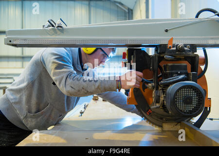 Ein junger Teenager-jungen Mann mit Ohr Schutz und Sicherheit Brille Sägen von Holz, die lernen, wie man sicher und sachgerecht verwenden eine macht sah in eine betriebliche Berufsausbildung Werkstatt, UK Stockfoto