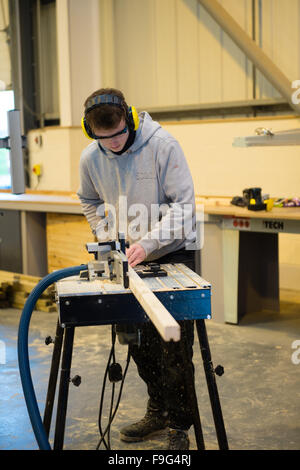 Ein junger Teenager-jungen Mann mit Ohr Schutz und Sicherheit Brille Sägen von Holz, die lernen, wie man sicher und sachgerecht verwenden eine macht sah in eine betriebliche Berufsausbildung Werkstatt, UK Stockfoto