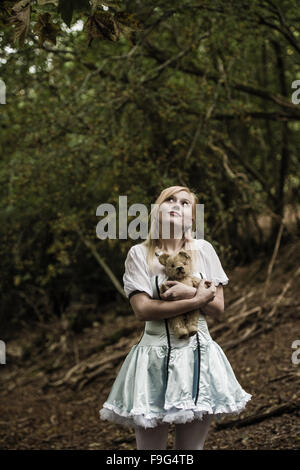 Malice in Wonderland / Albtraum: eine hübsche Blondine unschuldig aussehende junge Frau Mädchen tragen ein 'Alice' Art Kleid Kleid umklammert halten ihr Teddy Bär allein in einem verlassenen düsteren Wald Wald, UK Stockfoto