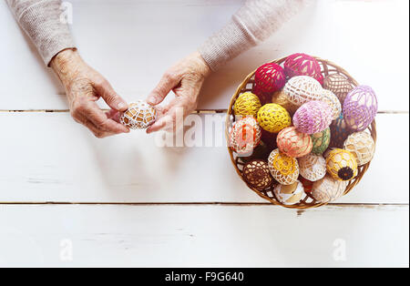 Händen der älteren Frau mit Korb voll von Ostern Eiern auf einem hölzernen weißen Hintergrund. Stockfoto