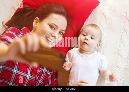 Niedliche kleine Baby-Mädchen und ihre Mutter nehmen Selfie auf einer Decke im Wohnzimmer. Stockfoto