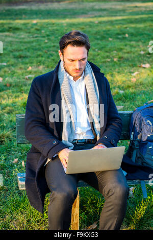 Schöne elegante Geschäftsmann sitzt auf einer Holzbank, arbeiten im Freien in einem städtischen Park, die Eingabe von Informationen auf seinem Laptop comp Stockfoto