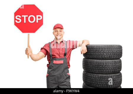 Junge männliche Mechaniker hält ein großes rotes Stoppschild und stützte sich auf einen Stapel von Reifen isoliert auf weißem Hintergrund Stockfoto