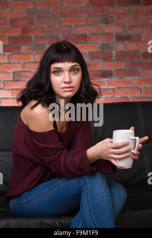 Charmante nachdenkliche junge Frau in Pullover und Jeans auf der Couch sitzen und trinken Tee über Ziegel Wand Hintergrund Stockfoto