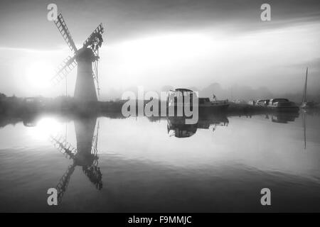 Atemberaubende Landschaft mit Windmühle und Fluss bei Sonnenaufgang am Sommermorgen in schwarz / weiß Stockfoto