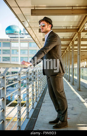 Voller Länge Portrait von Stylist junge Mann trägt Anzug und Hut auf der Seite von Balkon während stützte sich auf Handlauf in B Stockfoto