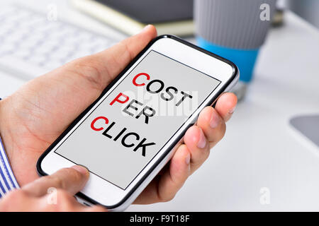 Nahaufnahme eines Geschäftsmannes, sitzt an seinem Schreibtisch mit einem Smartphone in den Händen mit den Text-Kosten pro Klick in seiner s geschrieben Stockfoto