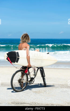 Brasilianische Surfer auf einem Fahrrad sitzen auf Fahrrad mit Blick auf die Wellen Ipanema Strand Rio de Janeiro Brasilien Stockfoto