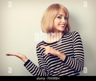 Schöne Make-up junge, blonde Frau hält und etwas in der Hand mit toothy Lächeln präsentieren. Getönten Porträt Stockfoto