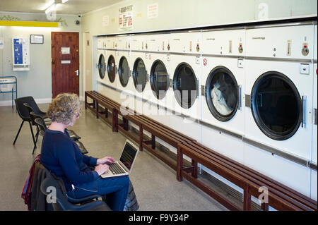 Frau, die im Waschsalon am Laptop arbeitet, während sie darauf wartet, dass die Laken trocknen Stockfoto