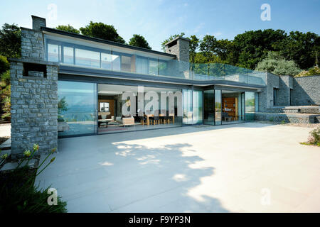 Von einem modernen Haus von der Terrasse auf den Hauptwohnbereich im Exterieur. Große Glas-Schiebetüren öffnen. Boden bis zur Decke Glasscheiben. Ein flaches Gebäude in einer Lage direkt am Meer. Stockfoto