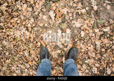 Männliche Füße in Blue Jeans und schwarze Schuhe stehen auf schmutzigen Waldboden mit herbstlichen Blättern, erste Person anzeigen Stockfoto