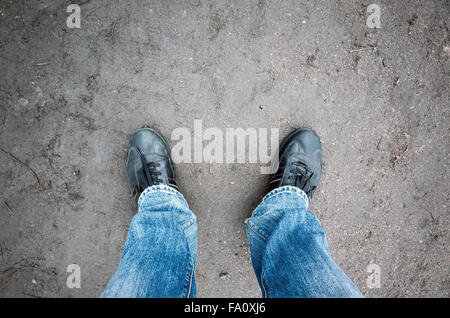 Männliche Füße in Blue Jeans und schwarze Schuhe stehen auf schmutzigen Landstraße, Egoperspektive Stockfoto