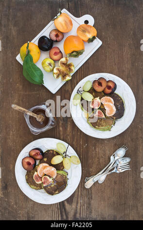 Frühstücks-Set. Selbstgemachte Zucchini-Pfannkuchen mit frischen Pflaumen, Mandarinen, Trauben, Feigen und Honig in den weißen keramischen Platten über rustikal Stockfoto