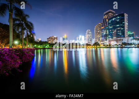 Moderne Wolkenkratzer und Palmen entlang Lake Rajada in der Nacht, im Benjakiti Park in Bangkok, Thailand. Stockfoto