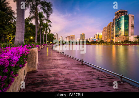 Moderne Wolkenkratzer, Blumen und Palmen entlang Lake Rajada bei Sonnenuntergang im Benjakiti Park in Bangkok, Thailand. Stockfoto