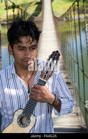 Junger Mann im gestreiften Shirt hält ein Charango, einem traditionellen Anden Saiteninstrument, hinter ihm eine Brücke über einem See Stockfoto