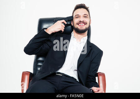 Lächeln auf den Lippen schön bärtigen Business-Mann im schwarzen Anzug und weißem Hemd sitzen im Bürostuhl mit Pistole an der Schläfe über weiß ba Stockfoto