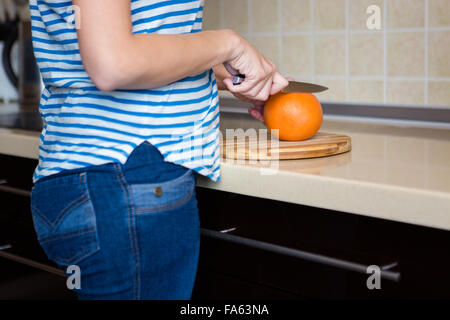 Schlanke junge Frau in gestreiften Oberteil und Jeans schneiden Grapefruit in Küche zu Hause Stockfoto