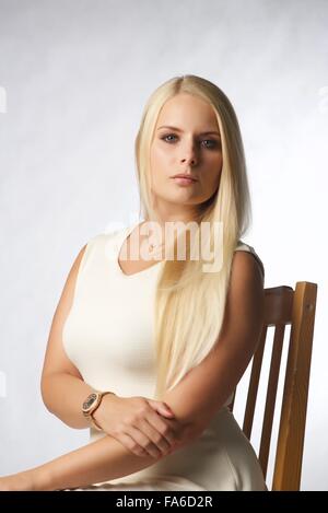 Ein blondes Mädchen sitzt auf einem Stuhl im Studio.  Sie trägt ein weißes Kleid und sucht nach Kamera Stockfoto