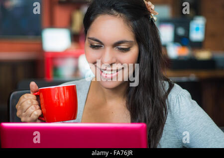 Attraktive Brünette trägt grauen Pullover sitzt am Tisch im Restaurant mit rosa Laptop arbeiten und halten rote Kaffeetasse schließen t Stockfoto