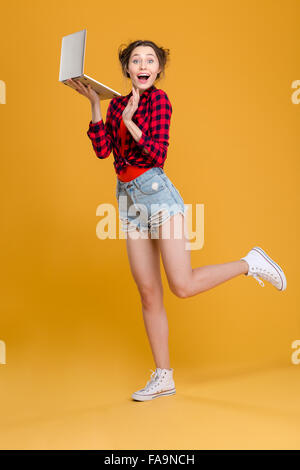 Gesamte Länge der süße schöne fröhliche junge Frau im karierten Hemd und Jeans Shorts posiert mit Laptop auf gelbem Hintergrund Stockfoto