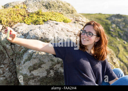 Junge Frau mit roten Haaren, wobei eine Selfie am Portmehor Point, Carn Galver, Cornwall, UK Stockfoto