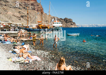 Santorini, Korfos, Aussicht auf schmalen Kieselstrand, mit Touristen und Sonnenanbeter paddeln und am Strand. Blaues Meer, kleine Boote und felsigen Klippen. Stockfoto