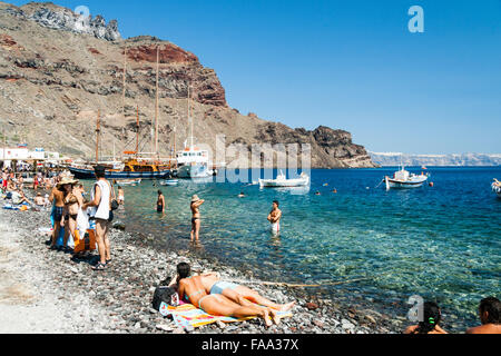 Santorini, Korfos, Aussicht auf schmalen Kieselstrand, mit Touristen und Sonnenanbeter paddeln und am Strand. Blaues Meer, kleine Boote und felsigen Klippen. Stockfoto