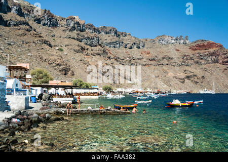 Santorini, Korfos, Aussicht auf schmalen Kieselstrand, mit Touristen und Sonnenanbeter paddeln und am Strand. Blaues Meer, kleine Boote und felsigen Klippen. Stockfoto