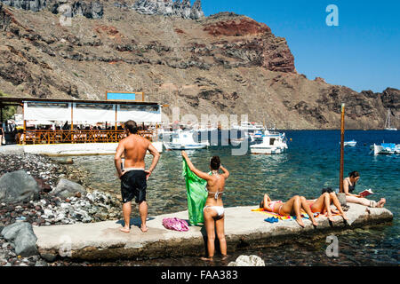 Santorini, Korfos, Aussicht auf schmalen Kieselstrand, mit Touristen und Sonnenanbeter paddeln und am Strand. Blaues Meer, kleine Boote und felsigen Klippen. Stockfoto